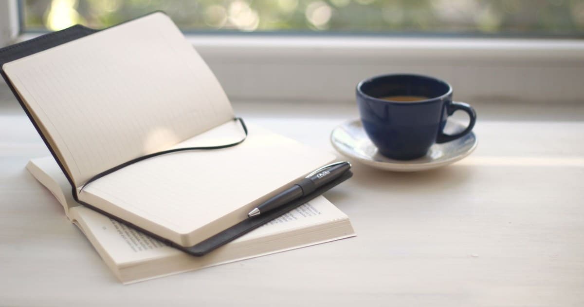 Cozy workspace setup with open notebook and coffee near a sunlit window.