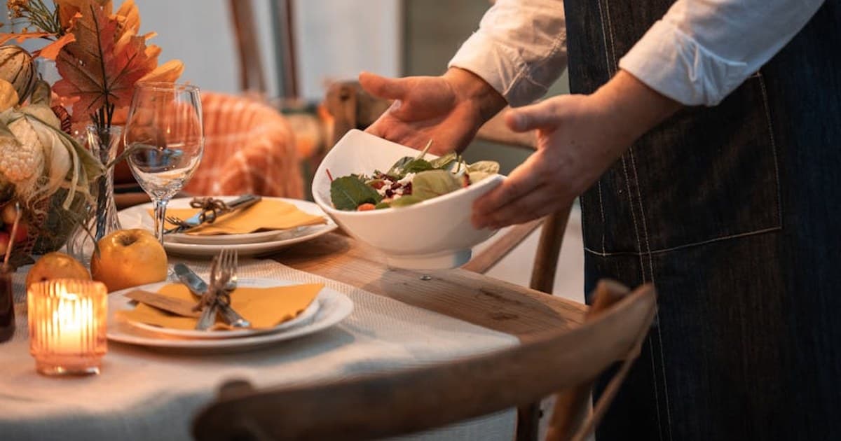 Warm and inviting autumn table setting with a person serving salad, perfect for Thanksgiving dinner.