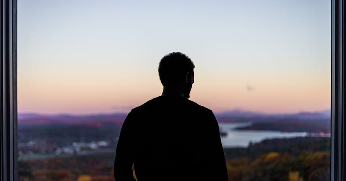 Man silhouetted against vibrant sunset, gazing out a window at a picturesque landscape with river and hills.