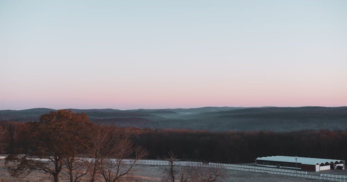 Serene rural landscape featuring bare trees, hills, and a barn at sunset.