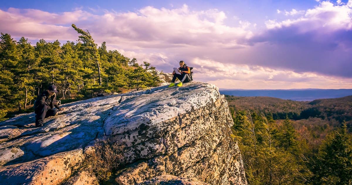 Hikers enjoy breathtaking views from a rocky cliff with vibrant skies and lush forests.