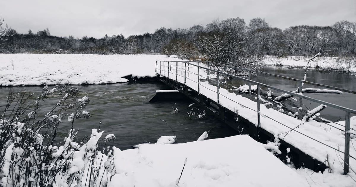A tranquil winter landscape featuring a snowy river with a metal bridge.